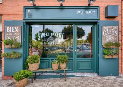 SWEET BAKERY, el obrador clásico inglés. En pleno barrio de Montequinto, siendo una antigua tienda de chucherías… Surge una imponente fachada verde carruaje completamente de madera y cristal, al más puro estilo ingles clásico. El diseño de un gran escaparate pintado a mano.Sweet Bakery es un pequeño obrador artesanal donde se elaboran grandes tartas, pasteles y cupcakes entre otros… Un diseño que destaca sobre la fachada de un edificio actual. Con puertas de hierro que se abren y adornadas con plantas y cestos de mimbre.Todo un concepto natural, artesanal y clásico con paredes de ladrillo antiguo, moldurones, zócalos. Un suelo hidráulico gastado con bancos y tapas de madera vieja, nos transporta 100 años atrás. Incluyendo una carpintería que nos permite ver el trabajo que se realiza de obrador en su interior.