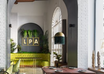 LPA, el nuevo restaurante de moda en Ayamonte, un proyecto de CM4. Con una gran parte de la cliente extranjera y siendo LPA uno de los referentes del municipio de Ayamonte (ciudad costera de la provincia de Huelva), el nuevo diseño e interiorismo de la reforma del local pretende sumergir al usuario en una experiencia fresca y acogedora, que realce los valores del clima mediterráneo.El primer obstáculo consistió en unir dos naves industriales y recuperar su estado y aspectos originales para después crear un espacio alegre y exótico, en el que la luz natural se derrama por todo el restaurante, y en la que los colores vivos y el blanco sirven de telón de fondo para una experiencia gastronómica inigualable.El local presenta dos zonas bien distinguidas, dos mundos. En el derecho nos encontramos con la zona de barra y coctelería, más informal, mientras que en la nave izquierda tenemos el comedor más grande. La cocina por otro lado, atraviesa las dos naves en la parte trasera, por lo que permite el paso de personal a las dos zonas de manera independiente.El ladrillo taco original de los muros, se pinta de blanco junto con el resto de paramentos para potenciar las grandes cristaleras y la luz natural tan presente en la costa. Con esto como base, se elige un mobiliario alegre, con tonos verdes claros y azules, que también podemos observar en el suelo de barro esmaltado. A esto se le suman un reservado decorado en tonos dorados y un salón superior desde el que se puede ver toda la altura de las naves y el sistema estructural de cerchas.Mármoles blancos y verdes, maderas, y vinilos en tonos claros completan los materiales bases del diseño.El objetivo y resultado final lleva al usuario a disfrutar de la comida como si al aire libre se tratase y acompañado siempre de un entorno mediterráneo.